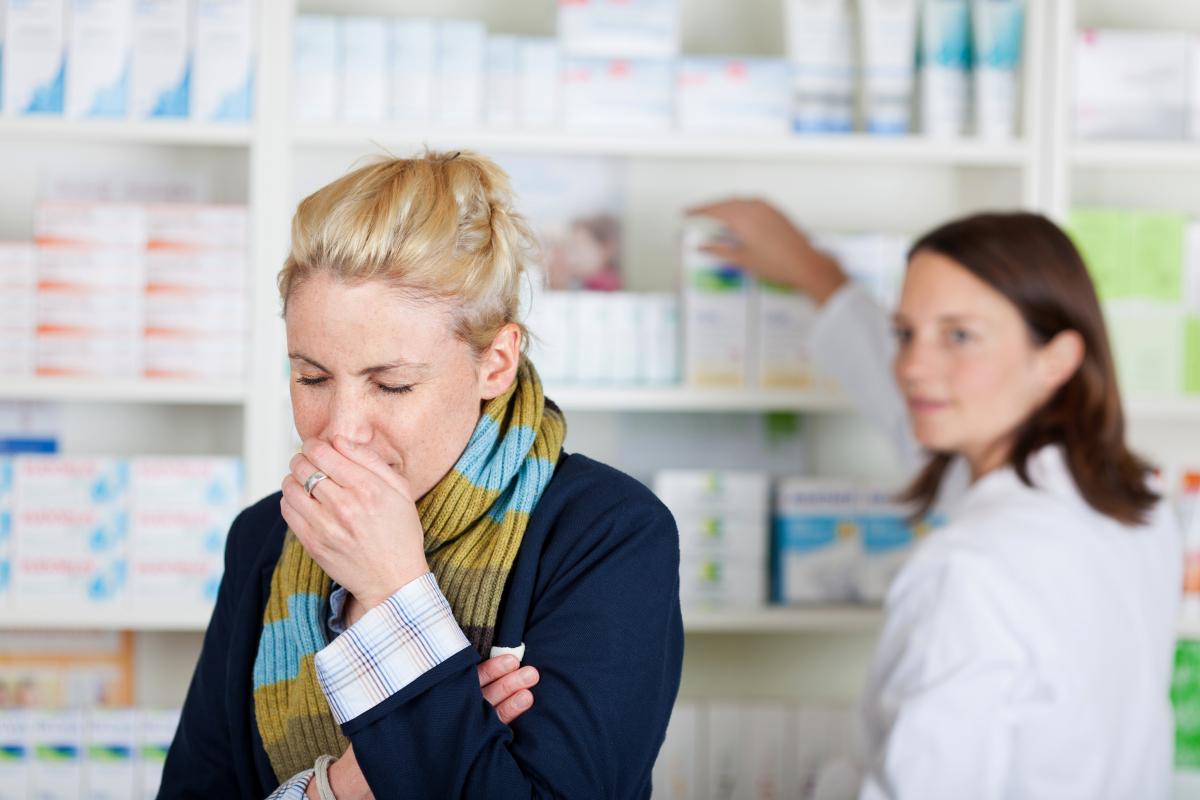 Kobieta stoi w aptece i kaszle, w tle farmaceutka sięga po lek z półki.