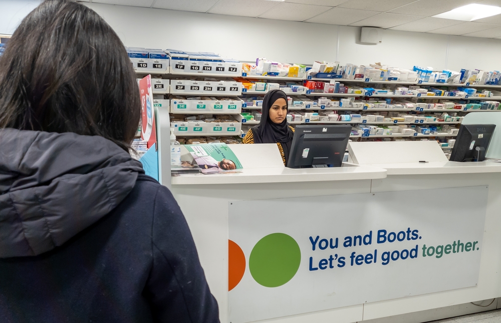 Wnętrze apteki w Londynie, pacjentka podchodzi do pierwszego stołu przy którym jest farmaceutka.